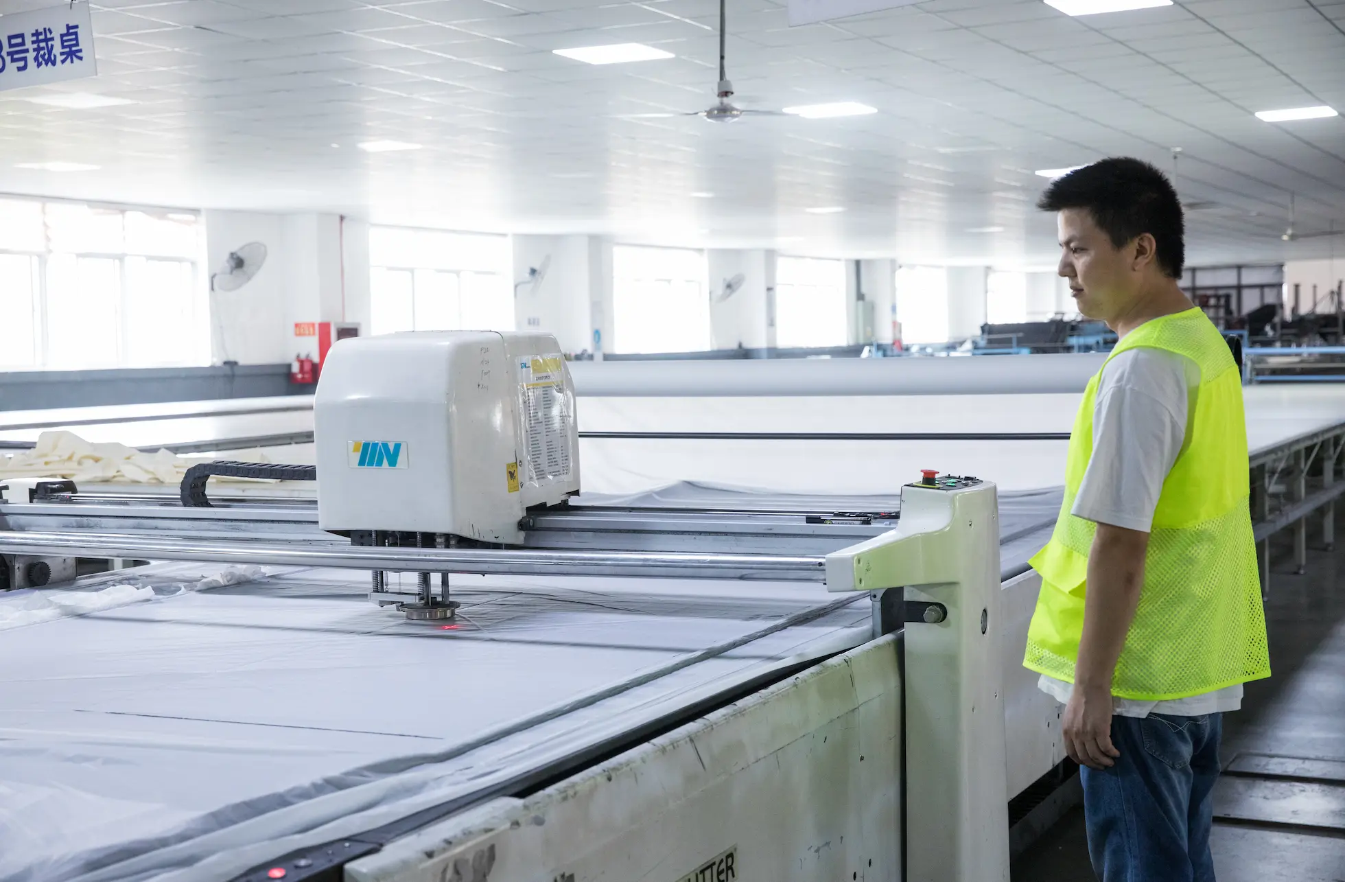 A factory employee supervises a large, automated machine that precisely cuts multiple layers of fabric for commercial umbrella canopies.