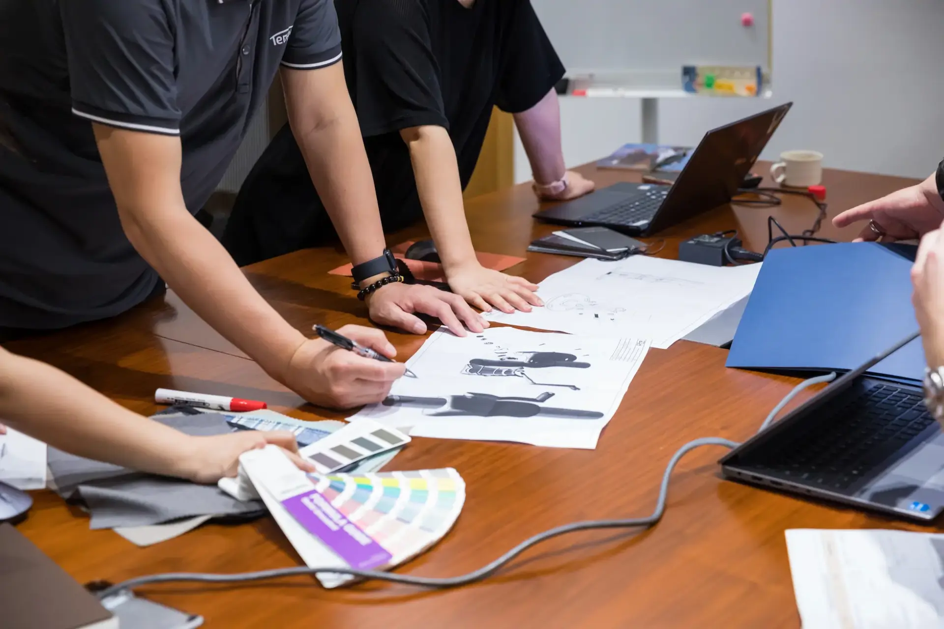 The product design team collaborates around a table, reviewing technical drawings and selecting materials and colors from swatches to develop a new outdoor umbrella concept.