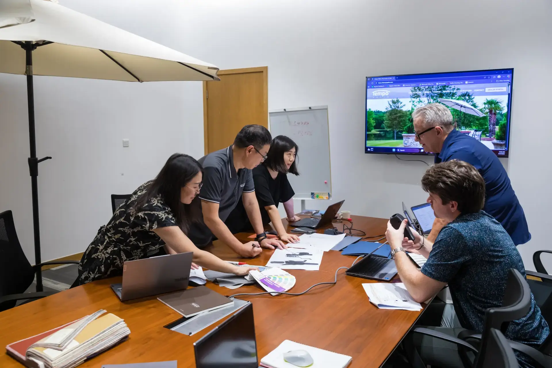 The product design team collaborates around a table, reviewing technical drawings and selecting materials and colors from swatches to develop a new outdoor umbrella concept.