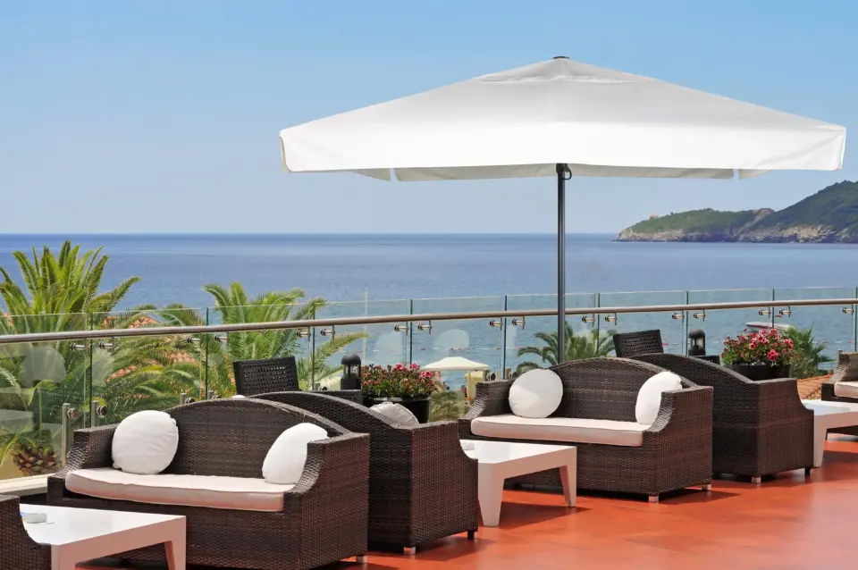 A large white Luciano patio umbrella providing elegant shade for wicker lounge furniture on a beautiful hotel terrace overlooking the ocean.