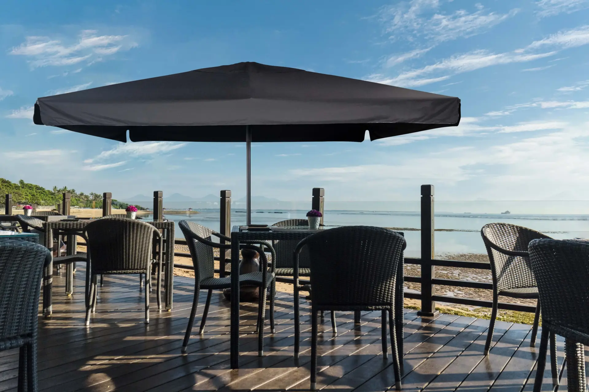A black Piazza center pole square market umbrella provides shade for a table and chairs on an outdoor wooden deck of a seaside restaurant.
