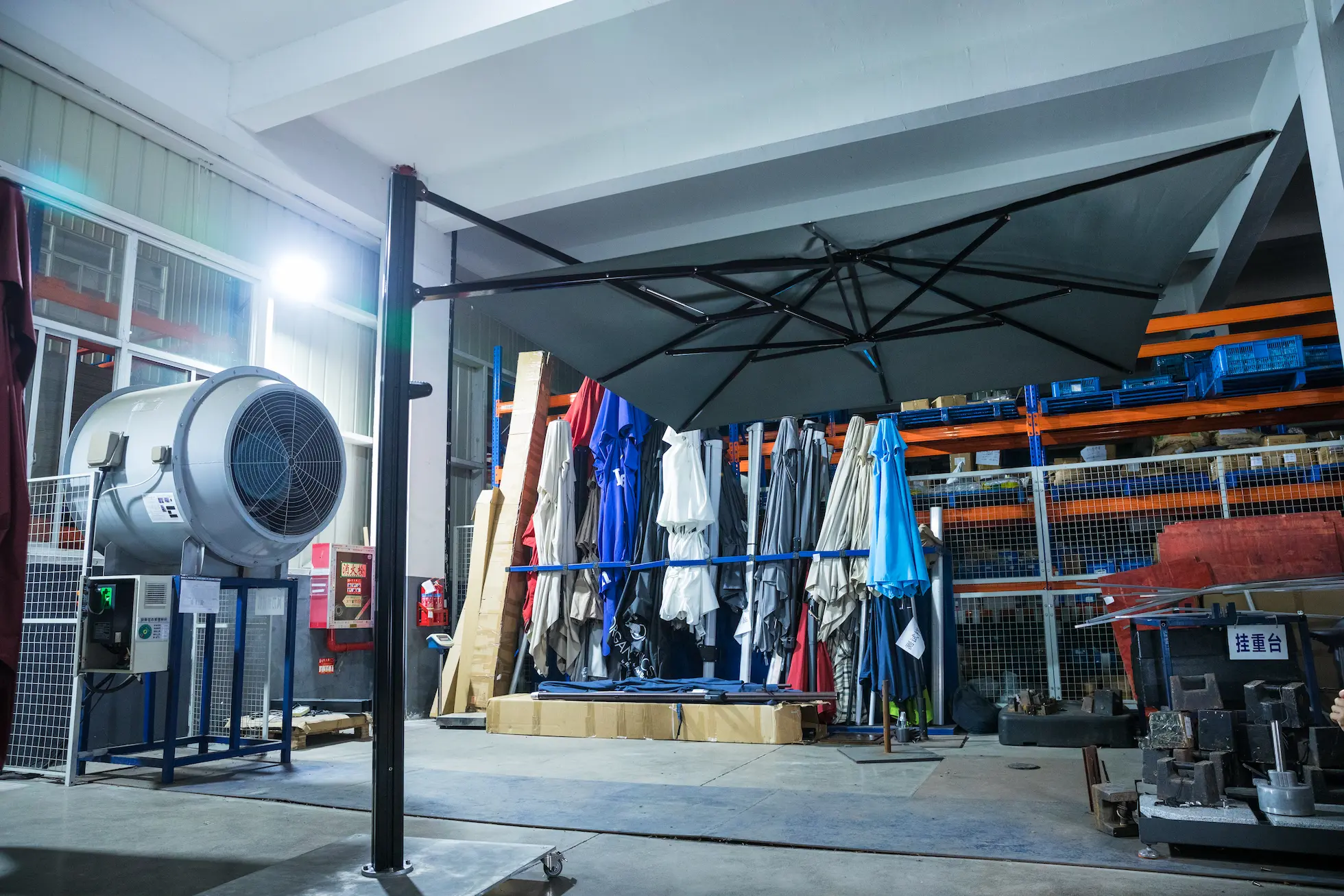 A fully assembled black cantilever umbrella stands in front of a large industrial fan for a wind resistance and stability test in the factory's quality assurance area.