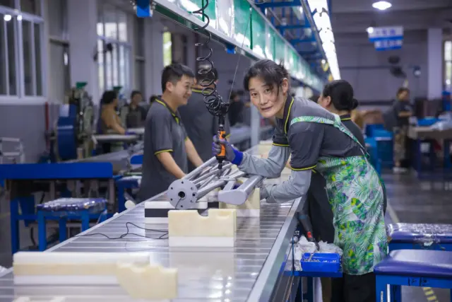 A smiling female worker skillfully assembles the intricate frame of a commercial outdoor umbrella on a busy and efficient production line.