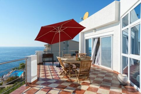 Tempo O2 umbrella in a red color, set up on a patio with a table and chairs, overlooking the sea.